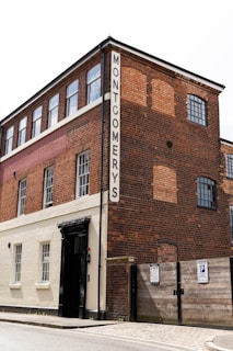 A three-story brick building with a sign labeled 'MONTGOMERYS' on a vertical panel. The building features large windows on each floor. The lower portion of the building is painted white and contains a black entrance door. There is a wooden gate with parking signs next to the entrance.