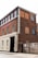 A three-story brick building with a sign labeled 'MONTGOMERYS' on a vertical panel. The building features large windows on each floor. The lower portion of the building is painted white and contains a black entrance door. There is a wooden gate with parking signs next to the entrance.