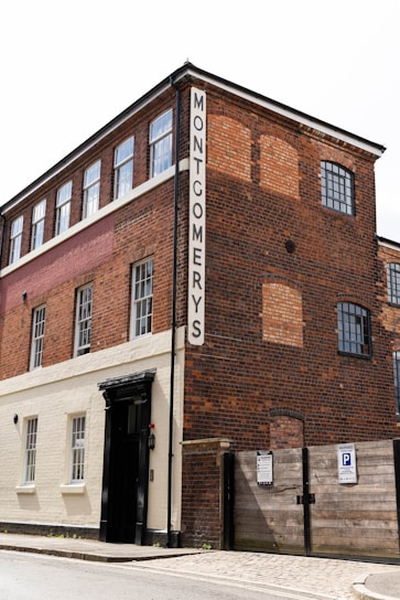 A three-story brick building with a sign labeled 'MONTGOMERYS' on a vertical panel. The building features large windows on each floor. The lower portion of the building is painted white and contains a black entrance door. There is a wooden gate with parking signs next to the entrance.