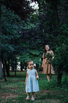A young child wearing a light blue dress stands in a lush, green forested area, surrounded by trees and grass. In the background, an adult woman in a brown dress is partially visible, appearing to watch over the child. The scene conveys a sense of tranquility and connection with nature.
