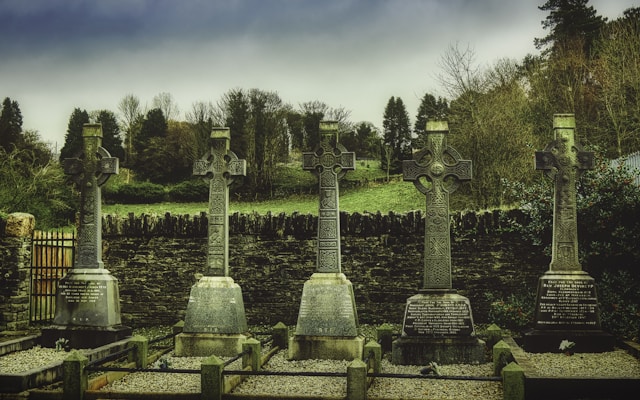 Four ornate Celtic crosses stand in a row, each serving as a gravestone in a serene graveyard setting. They are intricately carved and made of stone, positioned equally spaced on pea-gravel beds. Behind them, a stone wall separates the graveyard from a field dotted with trees, under a cloudy, overcast sky.