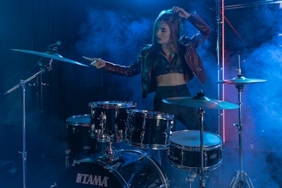 A woman stands confidently behind a drum set on a stage filled with blue and purple lighting and haze effects. She is wearing a dark leather jacket and a crop top, suggesting a rock or music performance setting. Her raised arm and focused expression convey a sense of energy and presence.