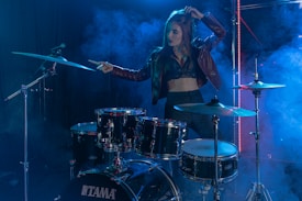 A woman stands confidently behind a drum set on a stage filled with blue and purple lighting and haze effects. She is wearing a dark leather jacket and a crop top, suggesting a rock or music performance setting. Her raised arm and focused expression convey a sense of energy and presence.