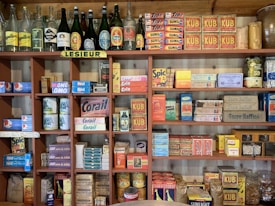 A vintage display of various retro packaging and bottles is arranged on wooden shelves. The shelves are filled with colorful boxes and cans of different food products, cleaning items, and glass bottles with old-style labels. The collection includes brands and labels primarily in French.