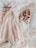 A delicate, cream-colored tulle dress paired with matching metallic pink ballet shoes. The soft textures and gentle hues create an elegant and serene atmosphere. The items rest on layered tulle fabric, enhancing the overall softness and femininity of the composition.