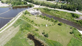 An aerial view of a lush green park with several picnic tables scattered across a spacious lawn. A river runs alongside the park, with a bridge crossing over it. Roads and a few buildings can be seen in the background. Dense trees and vegetation surround the area, giving it a serene and natural atmosphere.