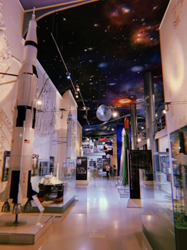 A museum exhibit featuring models of rockets and spacecraft with detailed space-themed decorations on the ceiling. The room is well-lit, showcasing various displays in glass cases, and there are informative signs and plaques visible around the area.