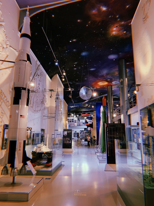 A museum exhibit featuring models of rockets and spacecraft with detailed space-themed decorations on the ceiling. The room is well-lit, showcasing various displays in glass cases, and there are informative signs and plaques visible around the area.