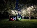 A nighttime outdoor setup with a beautifully lit tree adorned with string lights. Three bean bags in red, green, and blue are arranged around a small table on a grassy area, creating a cozy, inviting atmosphere. Surrounding foliage and distant small lights enhance the serene setting.