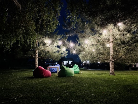 A nighttime outdoor setup with a beautifully lit tree adorned with string lights. Three bean bags in red, green, and blue are arranged around a small table on a grassy area, creating a cozy, inviting atmosphere. Surrounding foliage and distant small lights enhance the serene setting.