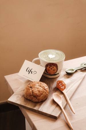 A cup of matcha latte with a heart design on top, placed on a wooden board beside a scone and a small dollop of jam. A wooden spoon and a card with the word 'life?' written on it accompany the setup. The cup features an owl design. The setting is cozy and simple.