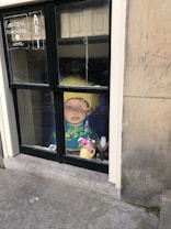 A window display features an artistic piece showing a portrait of a person with a yellow hat and detailed clothing. In front of the portrait, there's a vase with pink flowers and a quill. The window has handwritten elements, including a web address and the word 'ART', on the left pane. The building's exterior shows stonework with some signs of weathering.