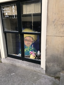 A window display features an artistic piece showing a portrait of a person with a yellow hat and detailed clothing. In front of the portrait, there's a vase with pink flowers and a quill. The window has handwritten elements, including a web address and the word 'ART', on the left pane. The building's exterior shows stonework with some signs of weathering.