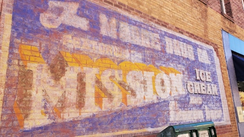 A vintage advertisement painted on a brick wall, featuring faded text promoting Mission Ice Cream. The lettering is large, with a mix of blue, white, and yellow colors, showing signs of wear and aging.