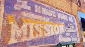 A vintage advertisement painted on a brick wall, featuring faded text promoting Mission Ice Cream. The lettering is large, with a mix of blue, white, and yellow colors, showing signs of wear and aging.