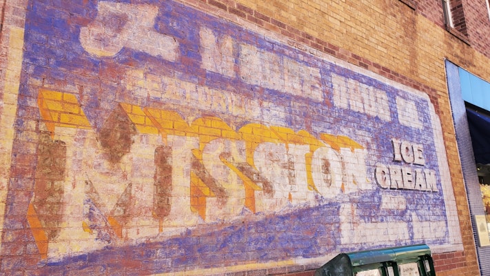 A vintage advertisement painted on a brick wall, featuring faded text promoting Mission Ice Cream. The lettering is large, with a mix of blue, white, and yellow colors, showing signs of wear and aging.
