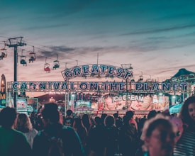 A vibrant carnival scene unfolds with colorful lights illuminating the entrance labeled 'Greatest Carnival on the Planet.' The sky is a mix of soft blues and pinks from the evening setting. Ice cream stands and various food stalls are visible, surrounded by a crowd of people enjoying the lively atmosphere. Overhead, a chairlift with several passengers adds to the festive ambiance.