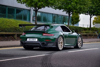 A green sports car with a rear spoiler is driving on a street lined with green trees and bushes. The car has a license plate number 803909 and features gold alloy wheels. In the background, there is a modern building with large windows and a sidewalk.