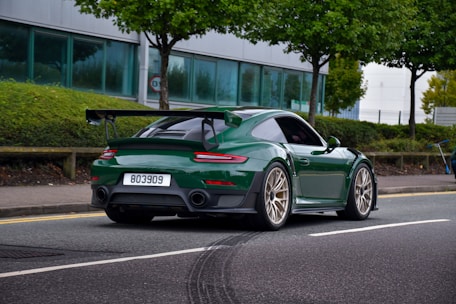 A green sports car with a rear spoiler is driving on a street lined with green trees and bushes. The car has a license plate number 803909 and features gold alloy wheels. In the background, there is a modern building with large windows and a sidewalk.