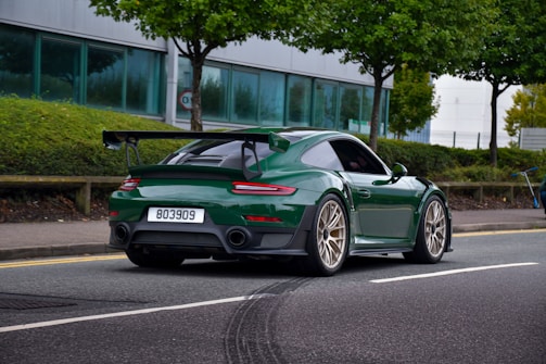 A green sports car with a rear spoiler is driving on a street lined with green trees and bushes. The car has a license plate number 803909 and features gold alloy wheels. In the background, there is a modern building with large windows and a sidewalk.