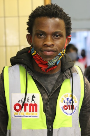 A person wearing a bright yellow safety vest with patches on the front. The person has short, curly hair and is wearing a colorful face mask with red, blue, and yellow patterns. They are standing in an indoor setting with tiled walls visible in the background.