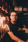 A person sits in a dimly lit environment with warm, ambient lighting creating a cozy atmosphere. The surroundings include reflective surfaces and glowing neon elements that add to the mood.