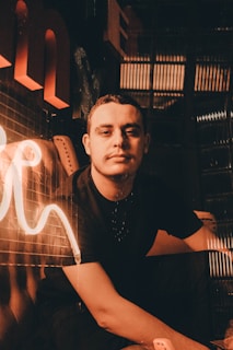 A person sits in a dimly lit environment with warm, ambient lighting creating a cozy atmosphere. The surroundings include reflective surfaces and glowing neon elements that add to the mood.