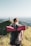 A person with a backpack and a red sleeping mat is walking along a grassy mountain ridge. In the distance, an observatory dome and a cross are visible against a backdrop of expansive views and a clear sky.