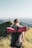 A person with a backpack and a red sleeping mat is walking along a grassy mountain ridge. In the distance, an observatory dome and a cross are visible against a backdrop of expansive views and a clear sky.
