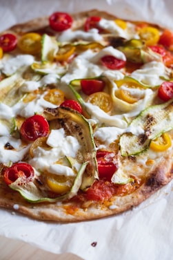 A freshly baked pizza topped with a variety of colorful ingredients, including cherry tomatoes in both red and yellow, thin slices of zucchini, and dollops of creamy white cheese. The pizza crust appears golden brown and slightly crispy.