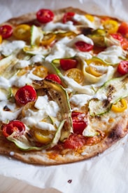 A freshly baked pizza topped with a variety of colorful ingredients, including cherry tomatoes in both red and yellow, thin slices of zucchini, and dollops of creamy white cheese. The pizza crust appears golden brown and slightly crispy.