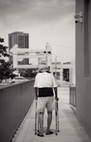 A person with mobility aids is walking along a sidewalk in an urban setting. The individual is using crutches and is dressed in casual summer clothing, including a hat and shorts. The walkway is flanked by railings, and in the background, there are tall buildings and structures. The image is in black and white, contributing to a somber or reflective mood.