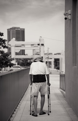 A person with mobility aids is walking along a sidewalk in an urban setting. The individual is using crutches and is dressed in casual summer clothing, including a hat and shorts. The walkway is flanked by railings, and in the background, there are tall buildings and structures. The image is in black and white, contributing to a somber or reflective mood.