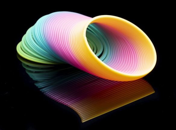 A colorful plastic spring toy, commonly known as a Slinky, with vibrant hues of yellow, pink, blue, and green, placed against a dark background. The smooth, wavy coils are neatly stacked and reflect on the surface below, creating a visually striking and harmonious pattern.