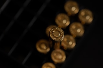 A close-up view of gold-colored bullet casings arranged in a box, with the headstamp reading '9mm Luger' visibly engraved on one of the casings. The lighting creates a reflective and metallic sheen on the surfaces.
