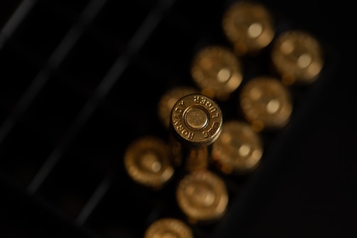 A close-up view of gold-colored bullet casings arranged in a box, with the headstamp reading '9mm Luger' visibly engraved on one of the casings. The lighting creates a reflective and metallic sheen on the surfaces.