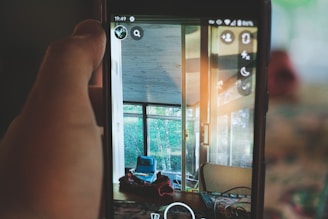 A hand holds a smartphone showing a photo of a room with large windows and visible greenery outside. Inside, there is a blue chair and some objects on the table near the window. The lighting is soft, creating a cozy ambiance.