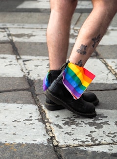 A person with tattooed legs is wearing black boots and standing on a paved surface. A rainbow flag is tucked into the boots, symbolizing LGBTQ+ pride.