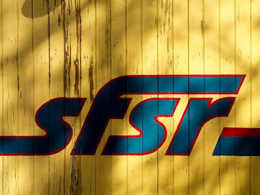 Faded yellow wooden boards serve as a backdrop for a large, bold and stylized blue and red logo that reads 'sfsr.' The wood appears weathered with some chips and visible nail marks, while shadows add texture and depth.