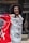A person with curly black hair and elaborate makeup is smiling and posing for the camera while holding a red and white banner. The banner features text in a bold script. The background shows an urban setting with a partially visible figure and building behind.