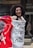 A person with curly black hair and elaborate makeup is smiling and posing for the camera while holding a red and white banner. The banner features text in a bold script. The background shows an urban setting with a partially visible figure and building behind.