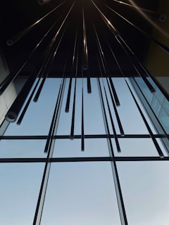 Long, slender tubes hang vertically from a ceiling, creating a modern and symmetrical composition. The tubes reflect light and appear metallic. The background features a large glass window allowing natural light to illuminate the space.