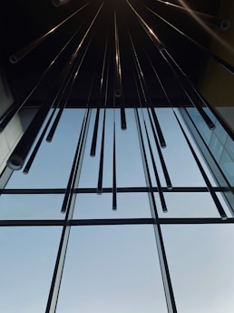 Long, slender tubes hang vertically from a ceiling, creating a modern and symmetrical composition. The tubes reflect light and appear metallic. The background features a large glass window allowing natural light to illuminate the space.