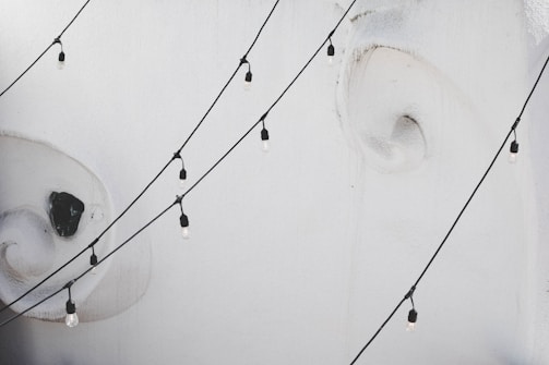 Several strings of small, clear, decorative lights are hanging diagonally against a white background. The wall behind the lights has subtle abstract designs in shades of grey and black, resembling brush strokes or swirls.