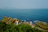Several people are exploring a rocky coastline, surrounded by lush green vegetation. The ocean stretches out into the horizon, with a few distant boats visible. The scene conveys a sense of calm exploration and leisure.