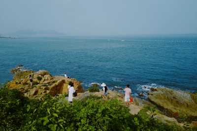 Several people are exploring a rocky coastline, surrounded by lush green vegetation. The ocean stretches out into the horizon, with a few distant boats visible. The scene conveys a sense of calm exploration and leisure.