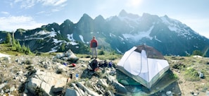 A hiker stands on rocky terrain overlooking a mountain range with patches of snow. A camping tent is pitched nearby, and various camping gear is scattered around on the ground. The scene is set in clear weather with bright daylight, providing a panoramic view of the rugged landscape.