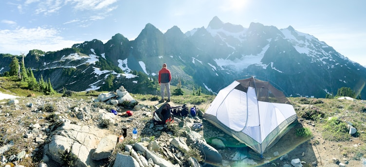 A hiker stands on rocky terrain overlooking a mountain range with patches of snow. A camping tent is pitched nearby, and various camping gear is scattered around on the ground. The scene is set in clear weather with bright daylight, providing a panoramic view of the rugged landscape.