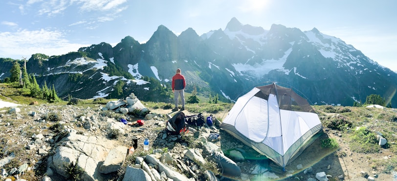 A hiker stands on rocky terrain overlooking a mountain range with patches of snow. A camping tent is pitched nearby, and various camping gear is scattered around on the ground. The scene is set in clear weather with bright daylight, providing a panoramic view of the rugged landscape.