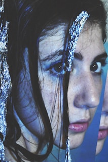 A close-up of a person's face with long dark hair partially covered by shiny metallic foil. The image is fragmented, creating a distorted and abstract look. The person has visible makeup and the focus is on their eyes and lips.